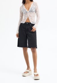 Cárdigan blanco de encaje de manga larga, pantalones cortos de mezclilla negros con dobladillos deshilachados y sandalias de plataforma beige con una correa en el dedo del pie.
