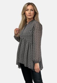 Czarna bluzka z długim rękawem w białe kwiatowe wzory, z plisowanym krojem, wysokim dekoltem i przezroczystymi rękawami, zestawiona z dopasowanymi czarnymi spodniami.