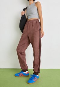 Hnědé teplákové kalhoty s černými pruhy na bocích; doplněné modrými teniskami s oranžovými detaily a šedým krátkým topem.