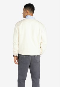 Sudadera de algodón blanca con mangas largas, cuello redondo, llevada sobre una camisa de cuello azul claro, combinada con pantalones gris oscuro.