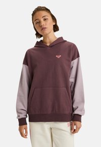 Mujer joven vistiendo una sudadera bicolor púrpura con un bolsillo frontal y pantalones blancos con un logo rojo en el muslo, de pie contra un fondo sencillo.