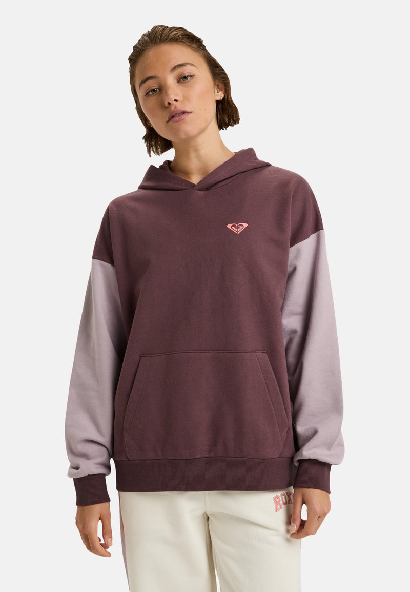 Mujer joven vistiendo una sudadera bicolor púrpura con un bolsillo frontal y pantalones blancos con un logo rojo en el muslo, de pie contra un fondo sencillo.