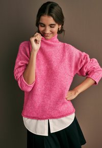 Maglione rosa lavorato a maglia con colletto alto, maniche ampie e una consistenza morbida, indossato sopra una camicia bianca con orlo spaccato.