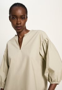 Frau mit geflochtenen Haaren, die eine beige, lockere Bluse mit drei Viertel langen, puffigen Ärmeln und einem V-Ausschnitt trägt, vor einem schlichten Hintergrund.