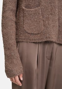 Nahaufnahme einer Hand neben einem strukturierten braunen Pullover mit einer Fronttasche und glatten, braunen, weiten Hosen.
