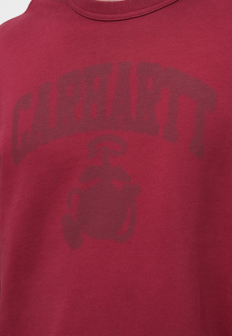T-shirt en coton marron foncé présentant un logo "Carhartt" surdimensionné en gris foncé et le contour d'une lanterne stylisée. Conception classique à col ras du cou.