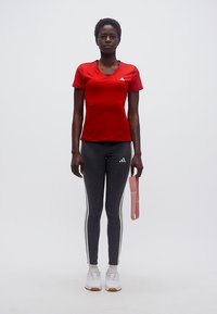 Γυναίκα με κόκκινο μπλουζάκι και γκρι κολάν κρατώντας μια ροζ λωρίδα γυμναστικής, στέκεται μπροστά σε λευκό απλό φόντο.