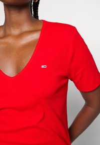 Punainen lyhythihainen V-pääntiäinen t-paita, joka on valmistettu ribbikudoksesta ja jossa on pieni broderattu logo sinisillä, punaisilla ja valkoisilla yksityiskohdilla.