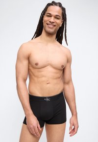 Lachende man met lange dreadlocks, gekleed in zwarte Calvin Klein boxershorts, staand tegen een effen witte achtergrond.