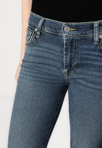 Jeans din denim albastru închis, cu un design ajustat, având un sistem de închidere cu nasturi, cinci buzunare și detalii de cusătură vizibile. Textura este ușor uzată.