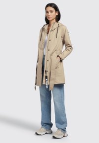 Beige waterproof jacket with a hood, adjustable drawstrings, snap buttons, quilted side pockets, and a logo accent on the tie.
