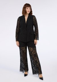 Zwart kanten pak met lange mouwen en wijde broekspijpen; heeft ingewikkelde bloemmotieven, een notch lapel en een gestructureerde stof. Zwarte hakken maken de look af.