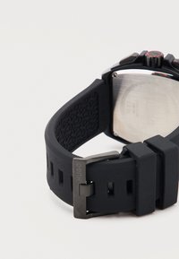 Μαύρο λαστιχένιο ρολόι με εγχάρακτη μεταλλική αγκράφα "HUGO" και ασημένιο μεταλλικό πίσω κάλυμμα ρολογιού σε λευκό φόντο.