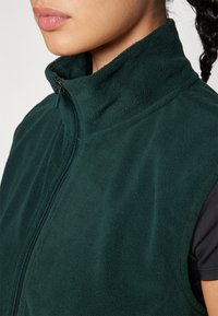 Chaleco de forro polar verde oscuro con cuello alto y cremallera frontal. Textura suave sin patrones ni adornos visibles. Vista en primer plano del área del cuello.