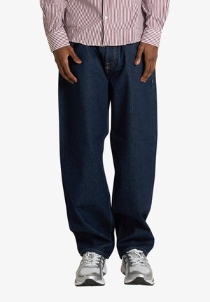 Persona che indossa jeans blu scuro a taglio largo, scarpe da ginnastica bianche e argento e una maglietta a maniche lunghe a righe rosse e bianche, in piedi con le mani lungo i fianchi.