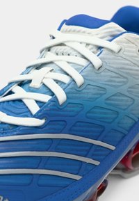 Pantof sportiv albastru și alb cu șireturi albe, panouri laterale texturate și crampoane roșii pe talpă.