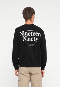 Black sweatshirt featuring large white text on the back: "Established in Nineteen Ninety" and "Designed by the J&J crew." Soft fabric.