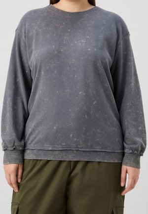 Personne portant un sweat-shirt à manches longues gris foncé délavé et un pantalon cargo vert olive, debout devant un fond clair uni.