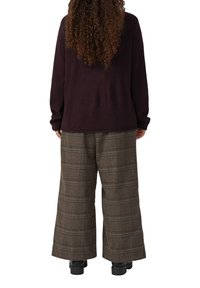Persoon met lang, krullend haar, gekleed in een donkerpaarse trui, brede bruine geruite broek en zwarte laarzen, staande met de rug naar de kijker gekeerd.