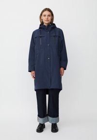 Cappotto blu navy, lungo fino al ginocchio, realizzato in tessuto intrecciato, con cappuccio, due tasche frontali e bottoni a pressione. Abbinato a jeans in denim scuro e stivali neri.