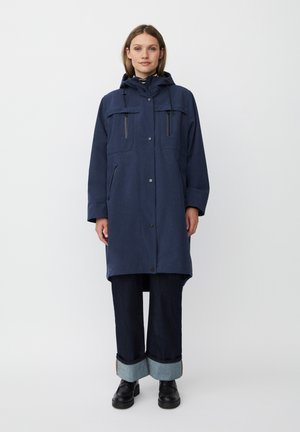 Cappotto blu navy, lungo fino al ginocchio, realizzato in tessuto intrecciato, con cappuccio, due tasche frontali e bottoni a pressione. Abbinato a jeans in denim scuro e stivali neri.