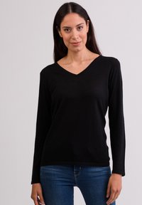 Schwarzes, langärmeliges V-Ausschnitt-Oberteil aus strukturiertem Stoff, mit lockerer Passform und Nahtdetail an den Manschetten, kombiniert mit blauen Jeans.