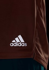 Camiseta atlética marrón con textura acanalada, que presenta un logo blanco de Adidas en la parte inferior izquierda. Mangas cortas y un ajuste holgado.