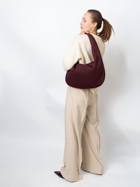 Vinröd quiltad axelväska med rundad form, med en texturerad yta och bred rem, dragen över en cremefärgad tröja och beige byxor.