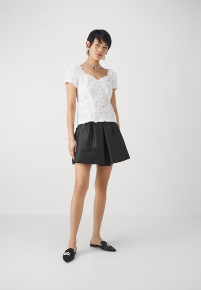 Weißes Spitzenoberteil mit floralen Mustern, kombiniert mit einem schwarzen Satinrock. Das Model trägt schwarze Mules mit silbernem Akzent und eine auffällige Halskette.