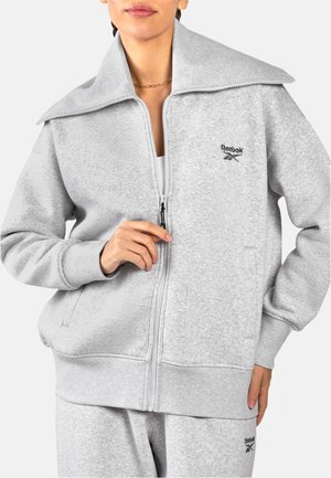 Femme portant un sweat-shirt zippé Reebok gris clair et un pantalon assorti, tenant la fermeture éclair près de la poitrine.