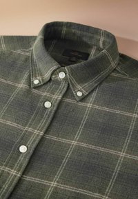 Camisa de franela verde con patrón a cuadros en verde oscuro y gris, con cuello abotonado y botones blancos. La etiqueta dice "Signature."