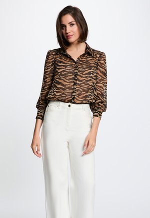 Kvinde med kort mørkt hår iført en brun og sort tigerprintbluse og hvide bukser med høj talje, stående mod en hvid baggrund.