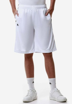 Uomo con pantaloncini sportivi bianchi con tasche, calzini bianchi con logo e scarpe da ginnastica bianche, in piedi con le mani nelle tasche.