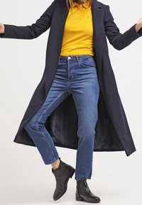 Lång marinblå kappa över en gul polo, bärs med blå raka jeans och svarta ankelstövlar. Släta texturer och minimal detaljer.