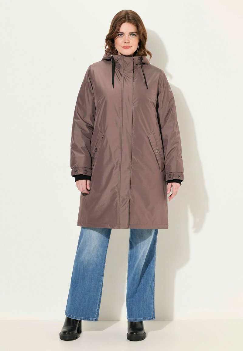 Brun vattentät parkas med hög krage, dragkedjestängning och justerbara dragsnören. Har sidofickor och svarta detaljer. Bärs över utsvängda jeans.