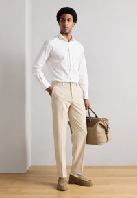 Camisa blanca de botones, pantalones beige y mocasines marrones. Sostiene una bolsa color canela con detalles marrones. Tejido suave y corte entallado.