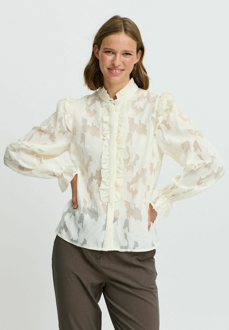 Blusa leggera color crema con colletto a volant, tessuto trasparente con un sottile motivo astratto e maniche lunghe a sbuffo. Abbinata a pantaloni scuri.
