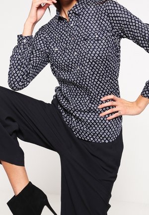 Kobieta ubrana w granatową koszulę zapinaną na guziki z nadrukiem, czarne spodnie o szerokich nogawkach oraz czarne czółenka na wysokim obcasie, pozująca z uniesionym kolanem.