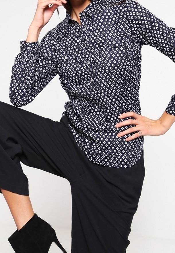 Femme portant une chemise bleu marine à motifs boutonnée, un pantalon noir à jambes larges et des bottines noires à talons hauts, posant avec un genou levé.