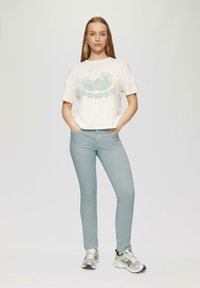 Witte cropped t-shirt met groene kers graphic en "POWER" tekst, gecombineerd met lichtblauwe strakke broek en zilveren sportschoenen.