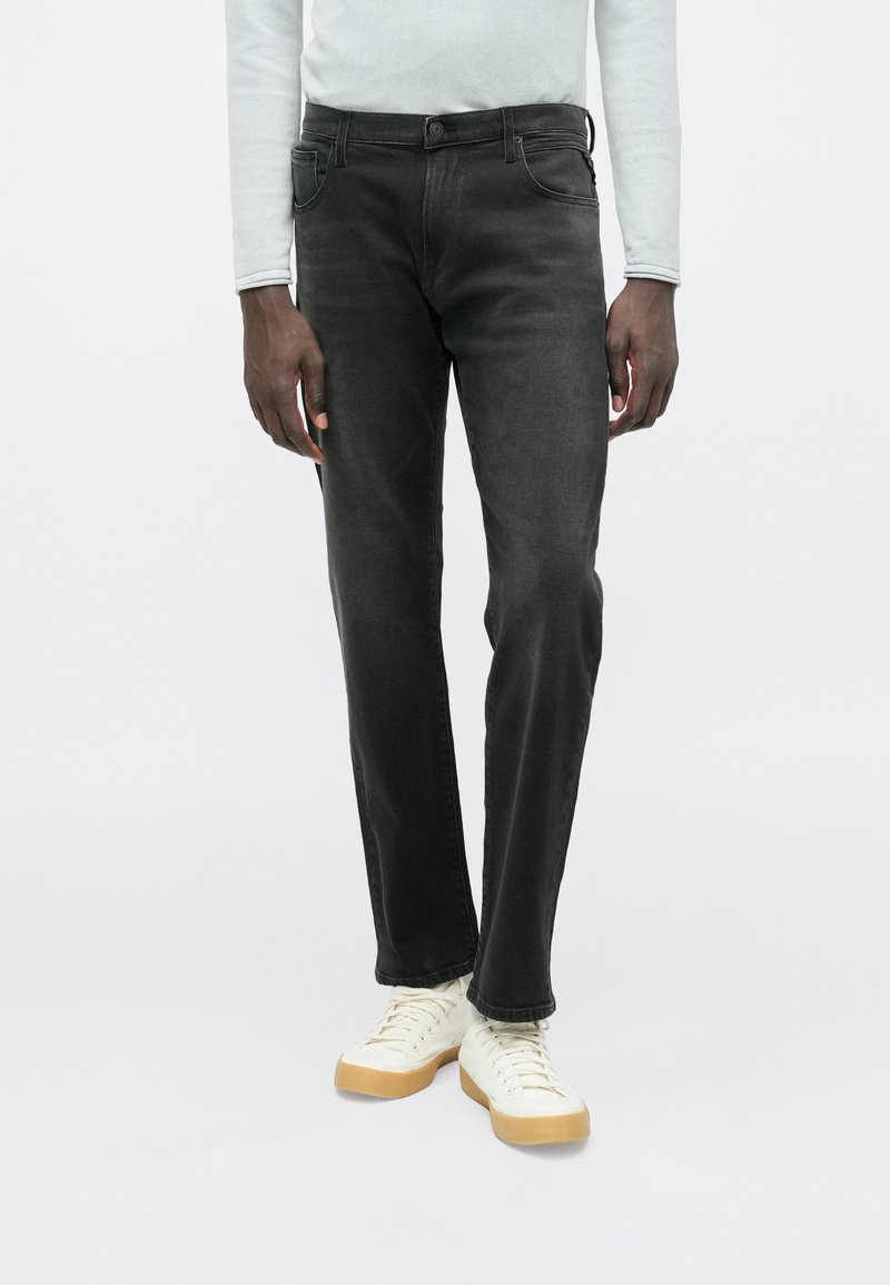 Μαύρο τζιν με ίσια γραμμή, πέντε τσέπες, ελαφριά ξεβάμματα και κλασικό κούμπωμα με κουμπιά, συνδυασμένο με λευκά αθλητικά παπούτσια και ανοιχτόχρωμο τοπ.