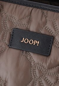 Vaatteetettu ruskea kangas, jossa on kohokuvioitu kukkaprintti, joka sisältää mustan tekstuurin etiketin, jossa lukee "JOOP!" kultaisella kirjotuksella.