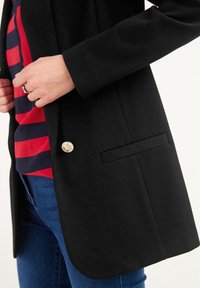 Henkilö, jolla on yllään musta bleiseri, jossa on kultainen nappi, punaraitainen ja siniraitainen paita, siniset farkut ja hopeasormus vasemmassa kädessä.