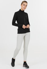 Zwarte sportjack met rits en lange mouwen, voorzien van een hoge kraag, gecombineerd met lichtgrijze leggings en zwarte sneakers.