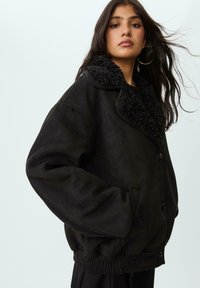 Crna zamšna jakna s plišanim, teksturiranim ovratnikom, prednjim gumbima i elastičnim rubom. Ima bočne džepove i opuštenu siluetu.