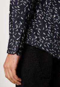 Tummansininen paita, jossa on valkoinen lehväkuvio, pitkät hihat ja napin hihansuut. Valmistettu pehmeästä kankaasta, jossa on hienovarainen kiilto ja vartalonmyötäinen malli.