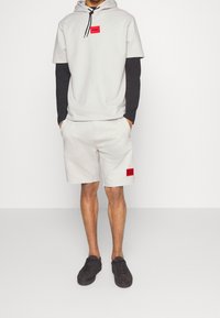 Sudadera con capucha gris claro con un parche de logo rojo, puesta sobre una camiseta de manga larga negra. Combinada con pantalones cortos gris claro con un parche de logo rojo. Zapatillas negras.