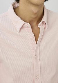 Chemise en velours côtelé rose clair avec col boutonné et texture côtelée verticale. Elle possède des boutons blancs et une coupe décontractée.