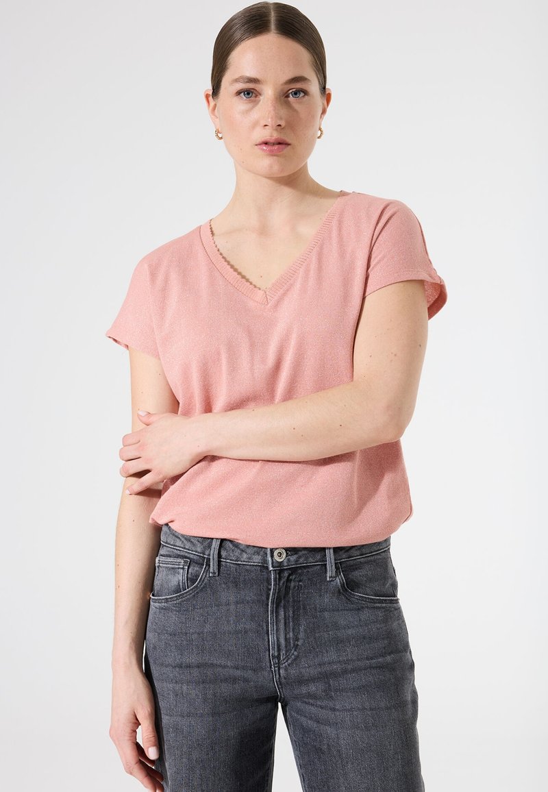 Haut rose à manches courtes et décolleté en V avec un léger éclat, associé à un jean gris foncé taille haute. Le tissu semble lisse et possède une coupe décontractée.