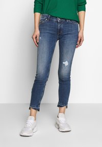 Pantalones vaqueros en un lavado intermedio con áreas desgastadas y un dobladillo ligeramente deshilachado, combinados con zapatillas deportivas blancas.